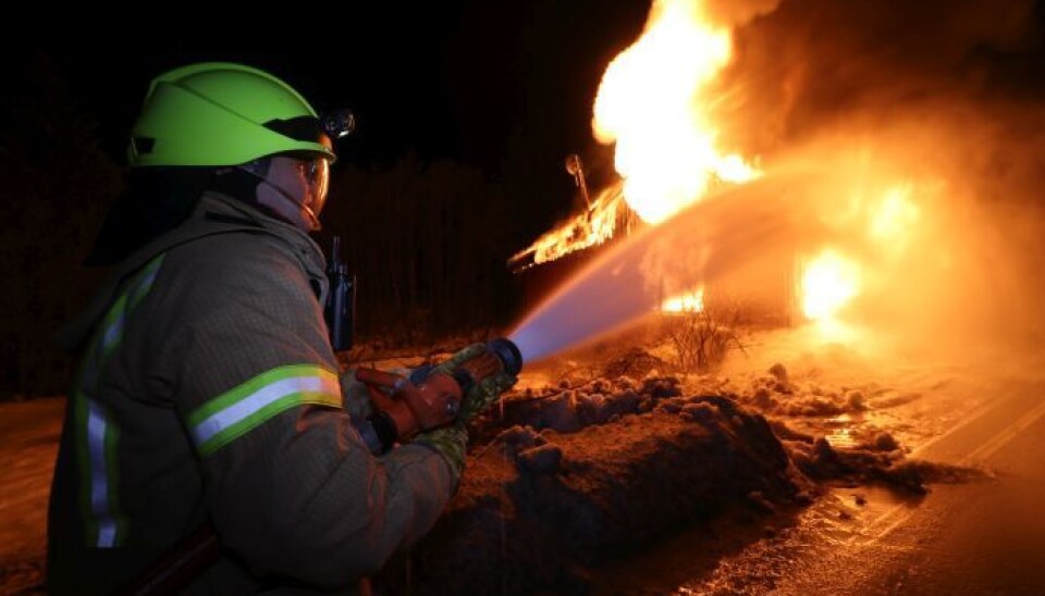 En brannmann i røykdykkerutstyr som holder en brannslange og spruter vann mot et brennende hus.