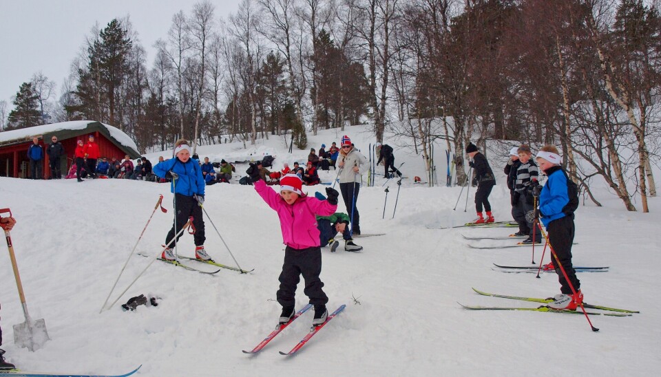 Flere barn som går og leker på ski. Mange mennekser og ei hytte i bakgrunnen.