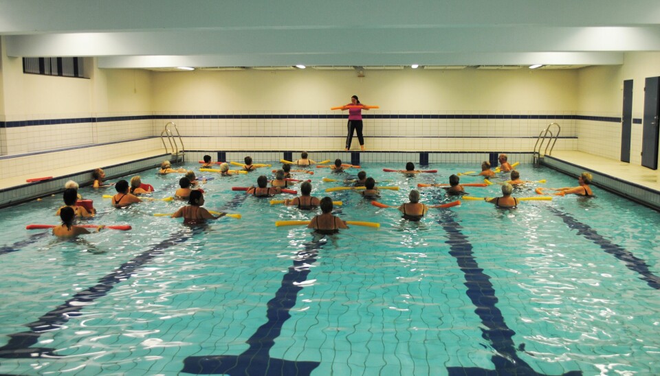 Mange mennesker som trener sammen i et basseng. Oppe på kanten står en instruktør.