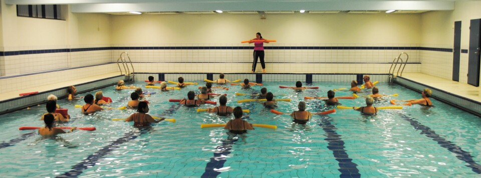 Mange mennesker som trener sammen i et basseng. Oppe på kanten står en instruktør.