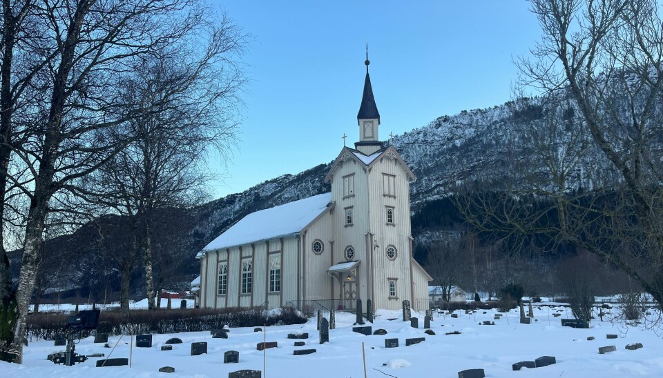 En kirke og en kirkegård, med fjell bak, trær, og snø på bakken.