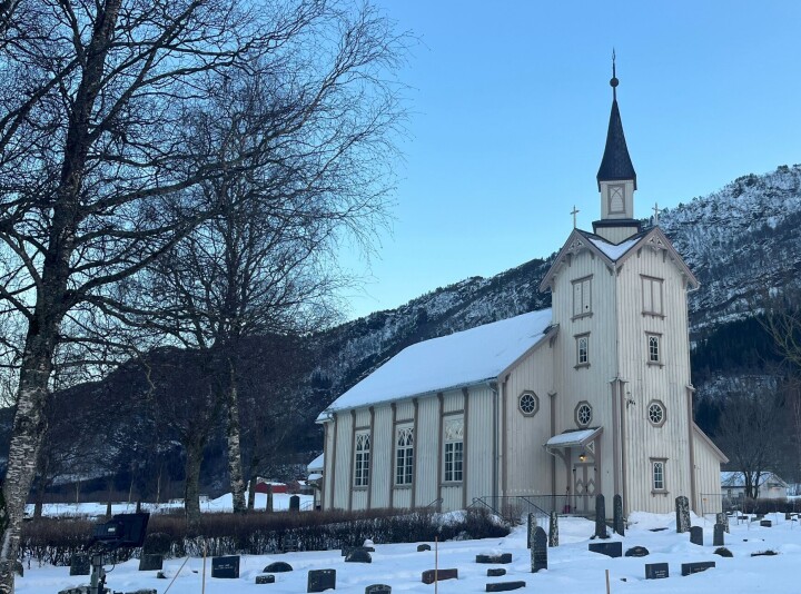 Ranes kyrkje En kirke og en kirkegård, med fjell bak, trær, og snø på bakken.