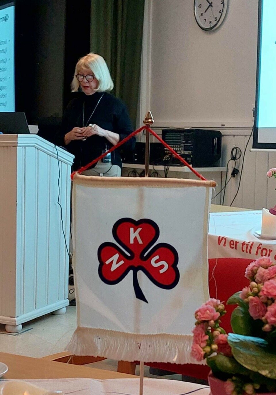 Elisabeth Swärd, seniorrådgiver i N.K.S Blond kvinne pater for forsamling med logoen til N.K.S. foran seg