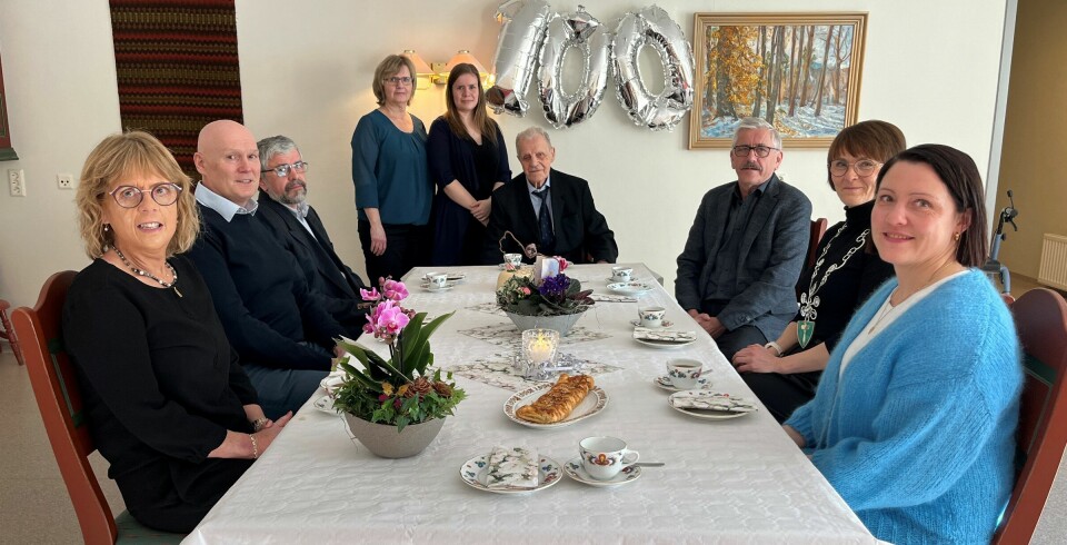 Ni smilende personer, menn og kvinner, sitter rundt et kaffebord. Ved enden av bordet sitter Johan Røen, som fyller 100 år. De andre er slektninger og gratulanter.
