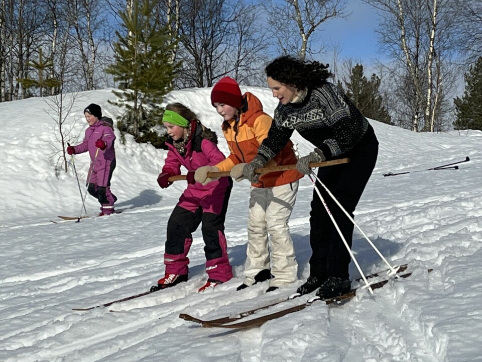 Tre jenter i vinterkjeledresser og ei dame i ullgenser kjører ski nedover en slak bakke i snøen. Tre av dem holder en gammeldags skistav i tre mellom seg.