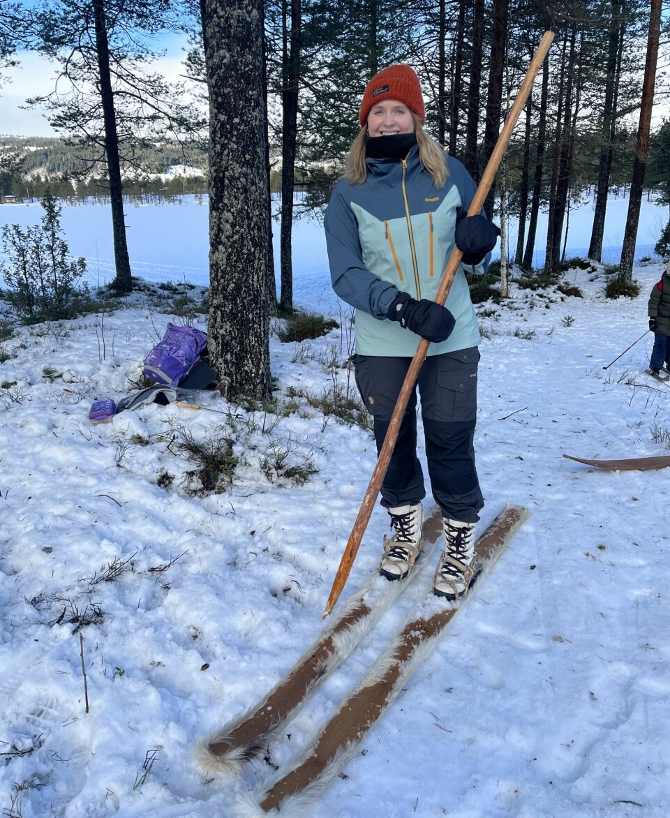 Ei smilende dame står i skogen med gammeldagse treski på beina. Under skia er det festet dyrepels. Hun holder en gammeldags lang skistav av tre i hendene.