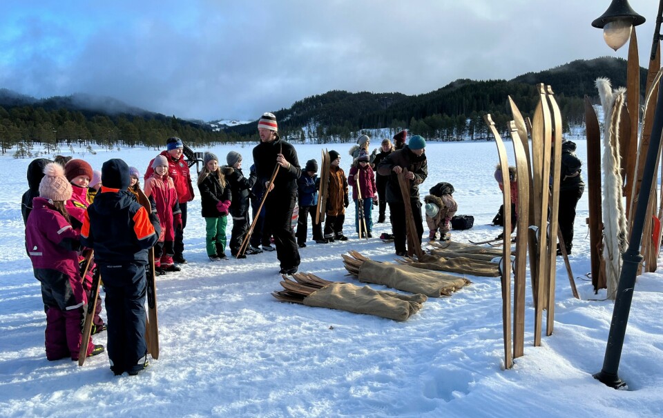 Mange barn og noen voksne i en halvsirkel i snøen rundt to menn som deler ut ski til dem. Flere par treski er plassert i snøen med tuppen oppover.