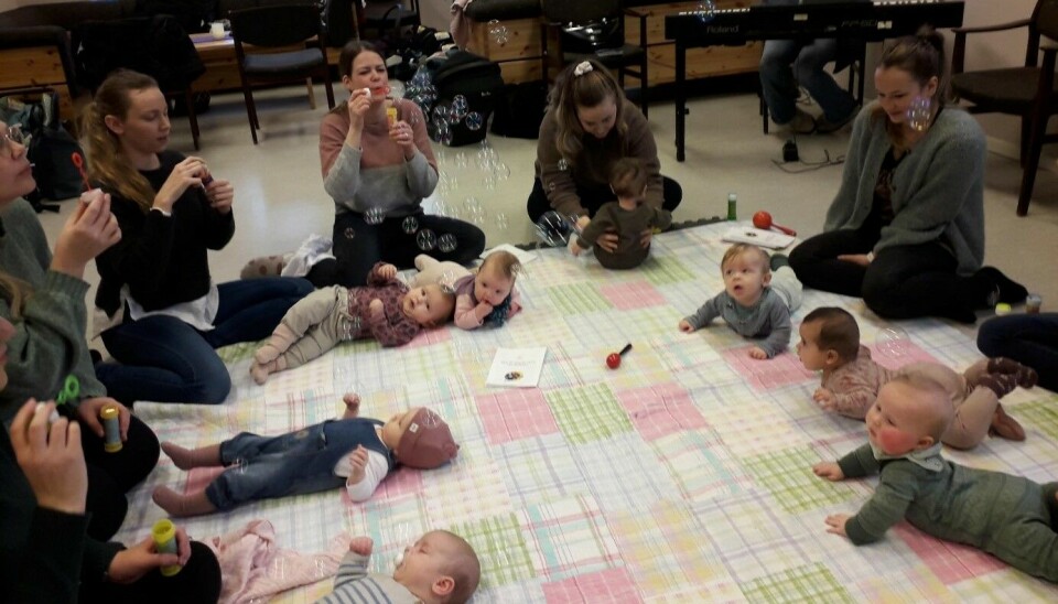 Flere babyer som ligger i ring på et teppe på gulvet. Mødrene sitter ved siden av.