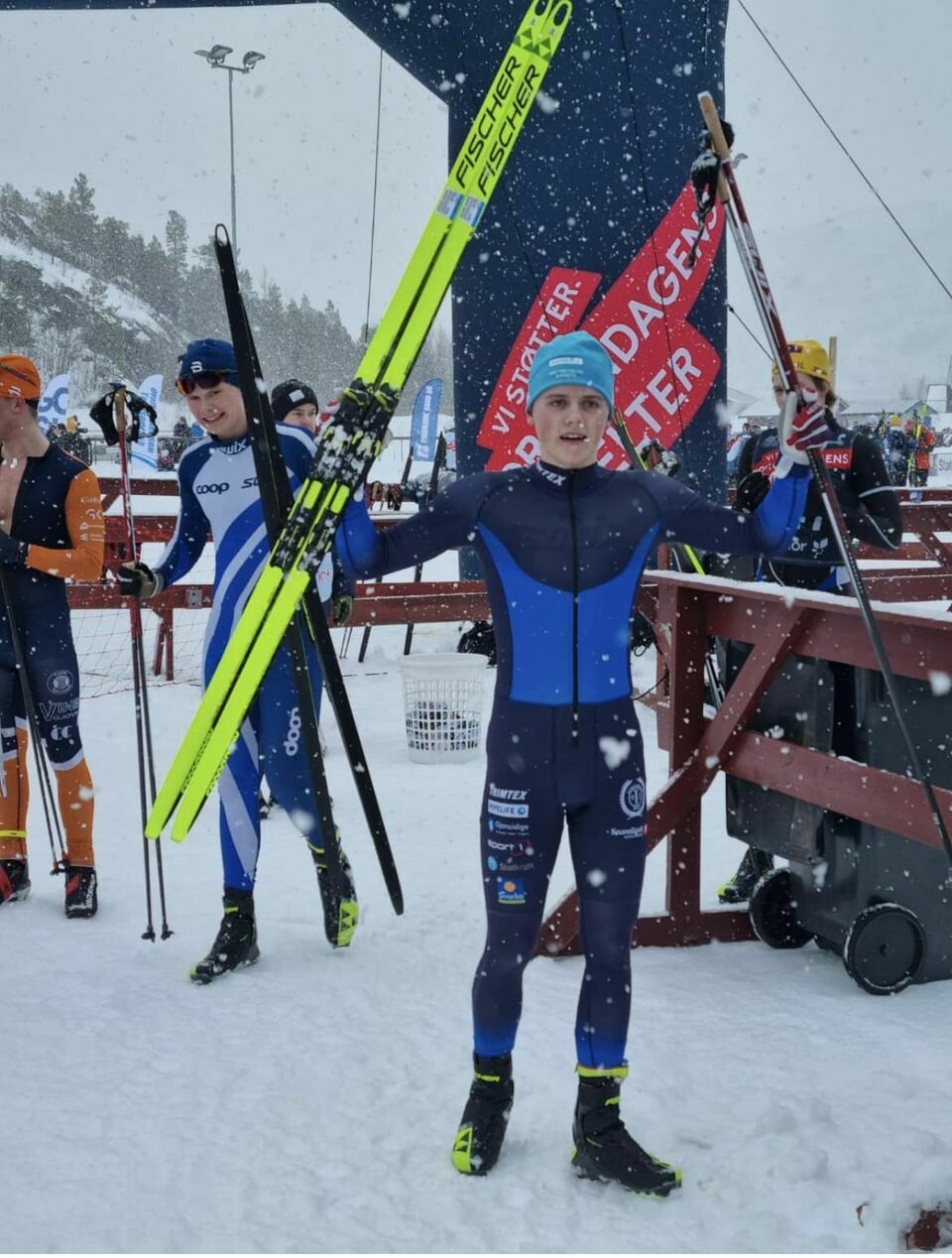 En ung mann i skidrakt som holder hendene i været. Han har et par ski i den ene hånda og stavene i den andre. Bak ser man flere skiløpener som er ferdig med sitt løp.
