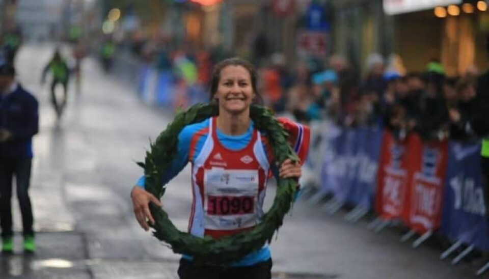 Kvinne med rød og blå løper og smiler med krans rundt halsen og det norske flagg bak seg.