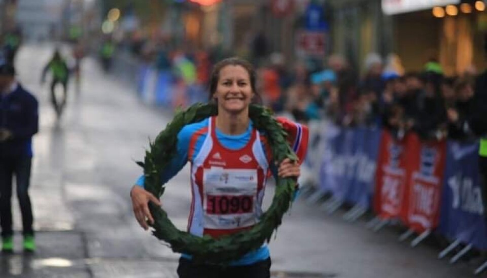 Bildet viser
Marianne Dønnem med laurbærkransen rundt halsen etter å ha vunne
kvinneklassa i Midnight sun maraton i Tromsø i 2018. Kvinne med rød og blå løper og smiler med krans rundt halsen og det norske flagg bak seg.