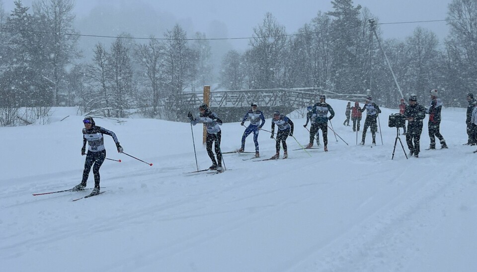 7 skiløpere ut fra start i skirenn. Det snør. Noen funksjonærer i bakgrunnen.