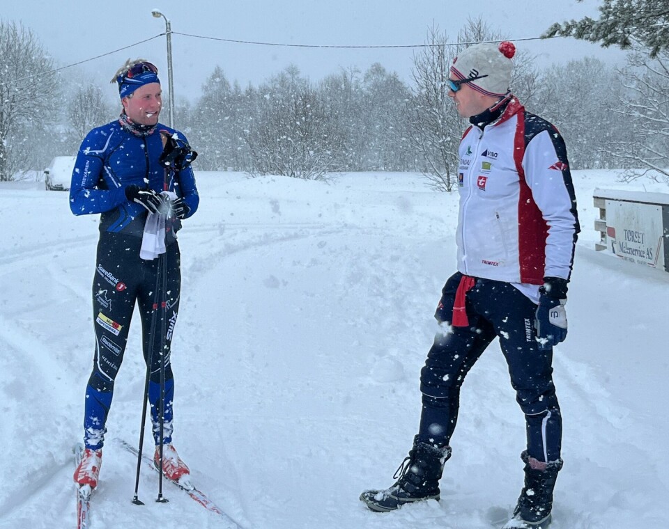 To menn som står og prater i snøen. Den ene har skidrakt og ski på beina, og stavene og et sammen foldet startnummer i hendene.