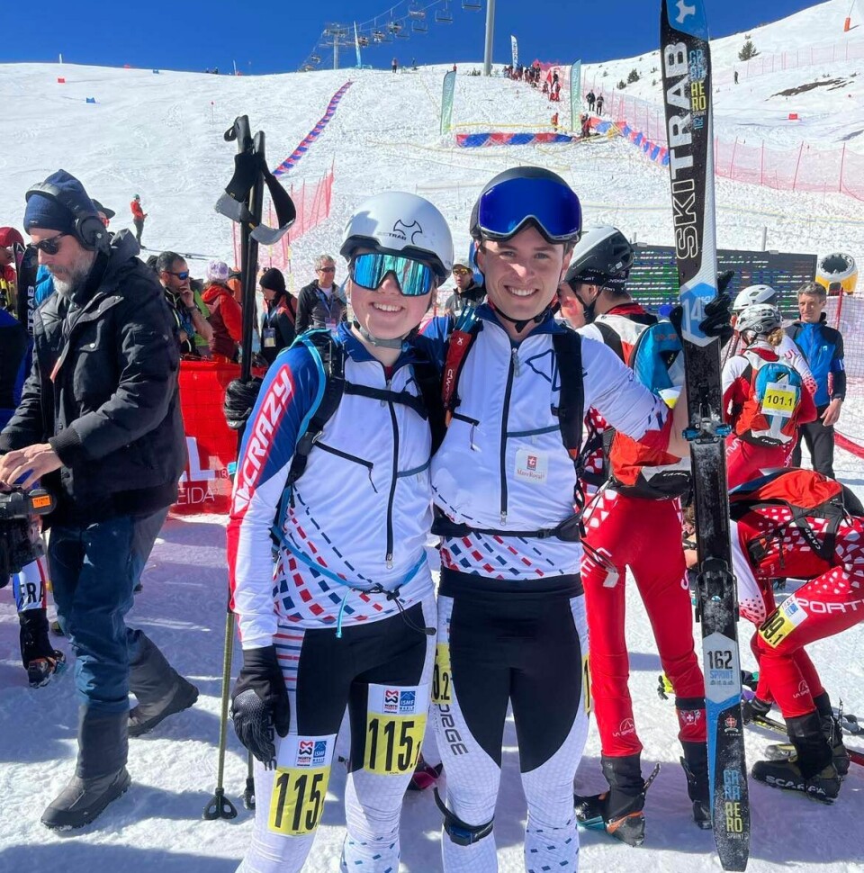 Ei ung jente og en ung mann i randonee-utstyr smiler mot kamera. Mannen løfter opp et par ski. Bak dem er en skibakke med skiheis, og flere utøvere og publikum.