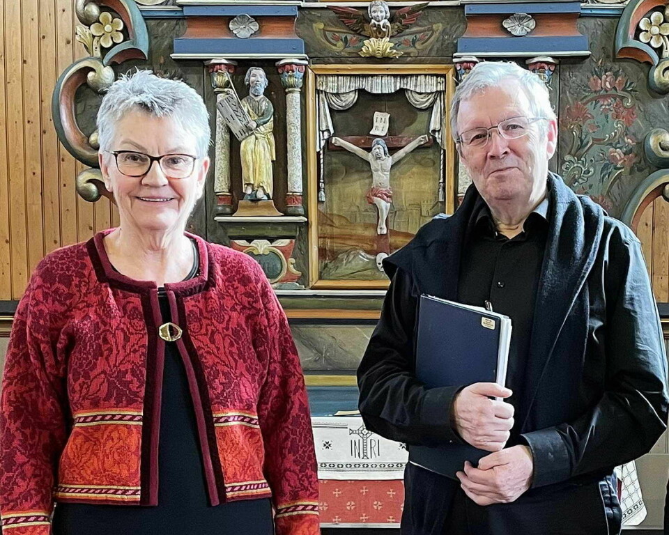 En kvinne og en mann, smilende, begge med grått hår og briller, foran en altertavle i ei kirke. Mannnen har ei skrivebok i hendene.