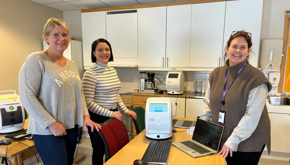 Tre smilende damer som står rundt et bord med en elektroniske medisindispenser