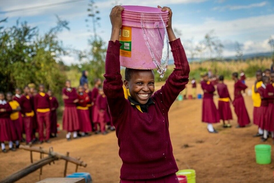 Bilde fra et utviklingsland. En smilende ungdom i skoleuniform med ei bøtte på hodet. Det renner litt rent vann ut fra bøtta.