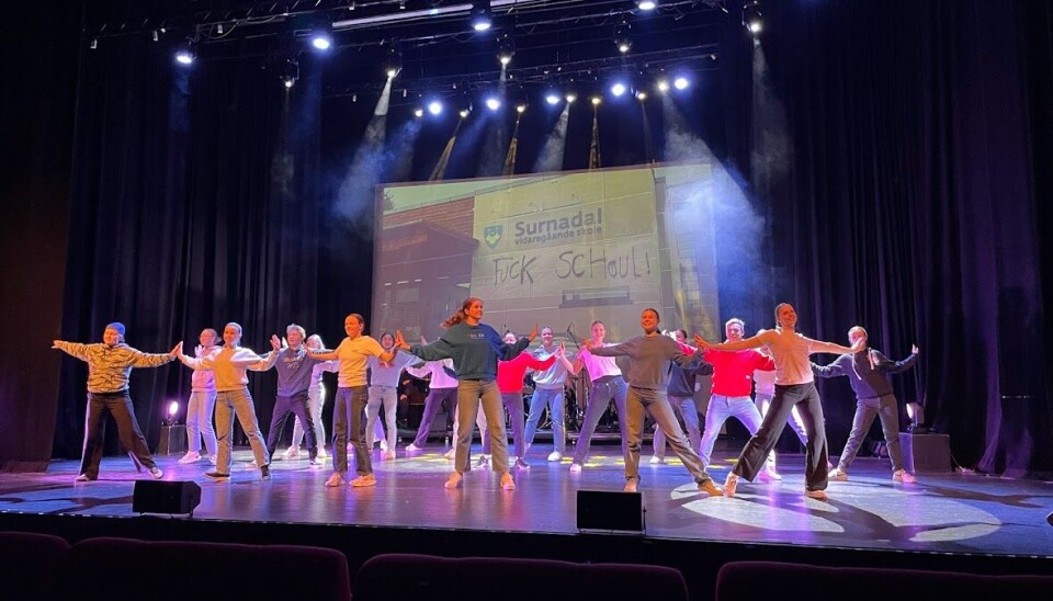 Mange ungdommer danser på scenen. På veggen bak dem et bilde av ytterveggen på Surnadal vidaregående skole, der det står 'Fuck Schoul' med spraymalt skrift.