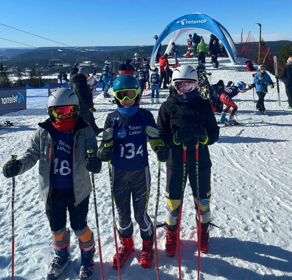Tre barn i alpinutstyr står ved siden av hverandre på en skiarena