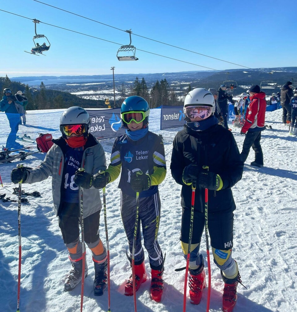 Tre barn i alpinutstyr står ved siden av hverandre ved en skiheis