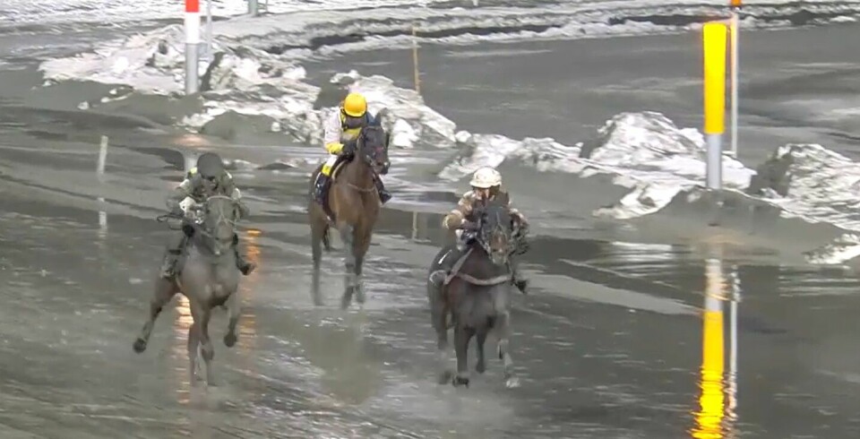 Tre hester med rytter i trav på løpsbanen. Det er vått på banen og litt snø.