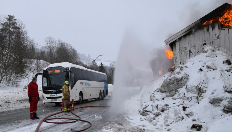En buss som kjører forbi et brennede hus. Mellom bussen og huset spruter det opp vann fra en brannslange, som beskytter trafikken mot varme og gnister.