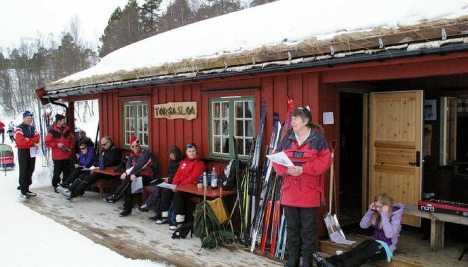 Mennesker står foran ei hytta som er dekt med snø og inntil hytteveggen står det ski lent og støtter seg på den.