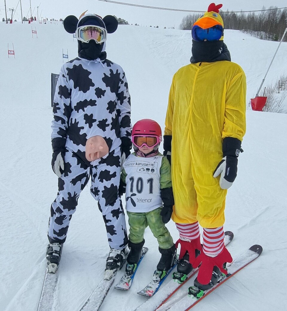 3 mennesker har akkurat kommet ned fra slalombakken. 2 voksne er kledd ut som ku og kylling, barnet i midten er kledd ut som drage.