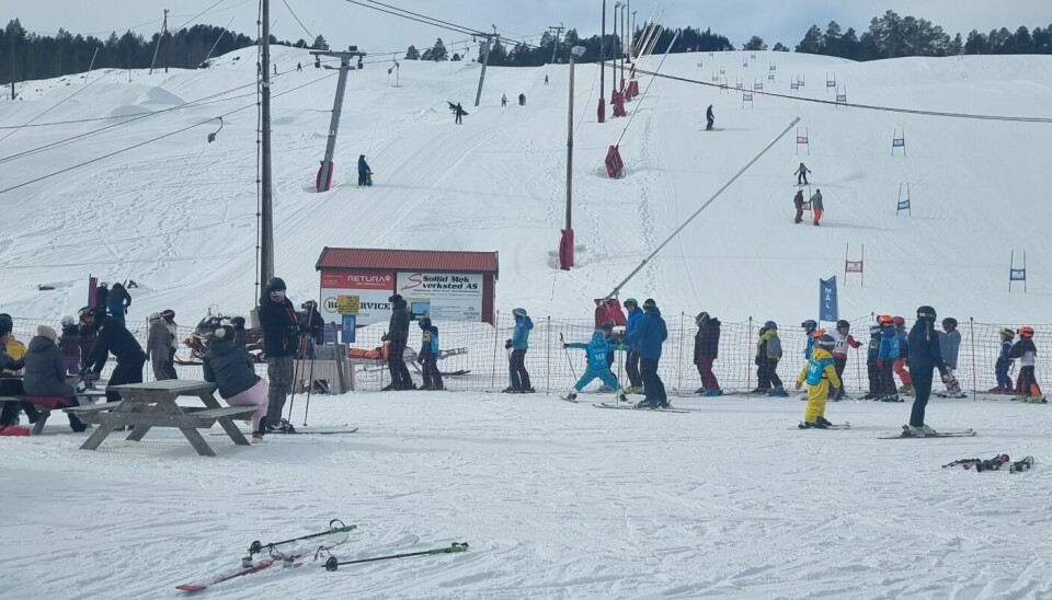 Mange barn og voksne står i kø til skiheisen. I bakgrunnen ser vi mennesker som kjører i bakken.