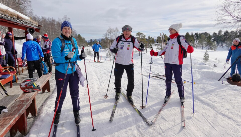 Tre voksne skiløpere i truklær klare for skitur. Komite og andre skiløpere i bakgrunnen.