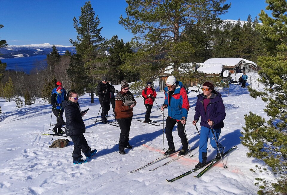 Flere mennesker på ski samla i fint vintervær med snø og sol. Furutrær og noen seterhus i bakgrunnen.