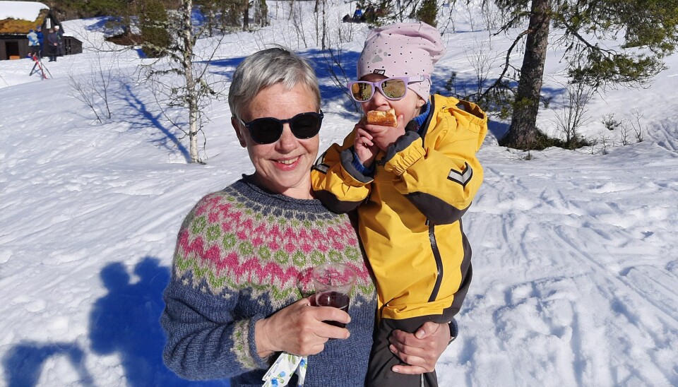 Ei smilende dame med kort hår, solbriller og ullgenser holder et lite barn på armen. Barnet har vinterdress, lue og solbriller. Fint vintervær med sol.