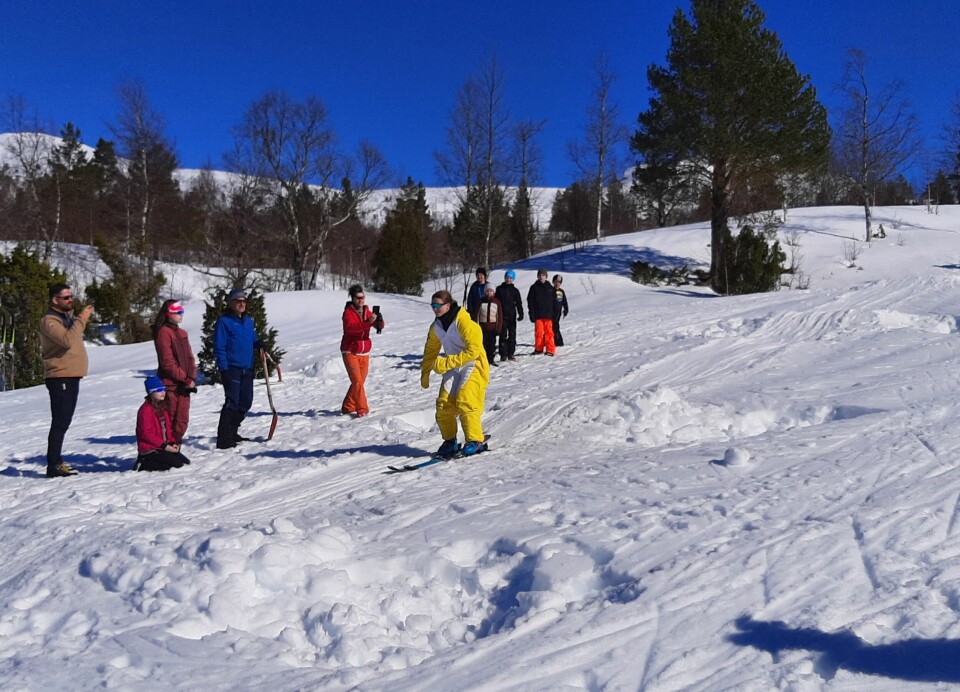 Ei jente i påskekylling-kostyme kjører på ski nedover ei slak kuleløype, blant mange andre mennesker, som er samla i fint vintervær med sol.