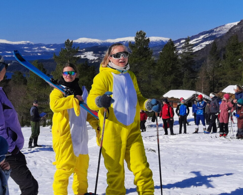To jenter i påskekylling-kostymer går på ski blant mange andre mennesker, som er samla i en skibakke i fint vintervær med sol.