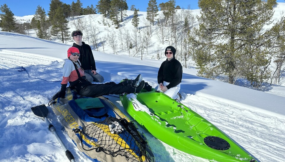 Tre ungdommer som sitter i snøen i en skibakke med en kajakk og en gummibåt.