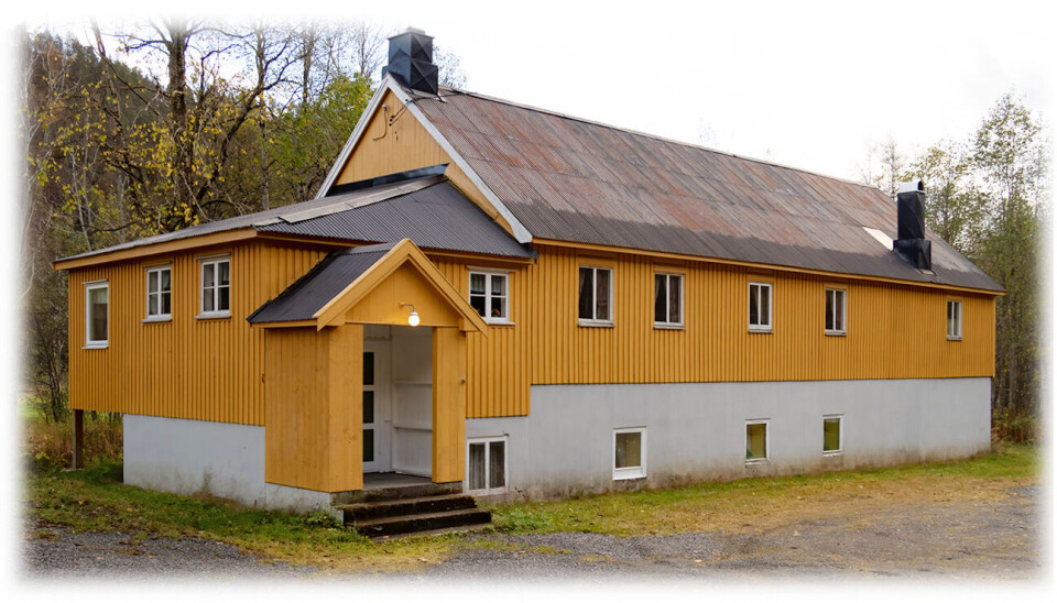 Et gult hus som har hvit mue nederst. Det er skog bak huset.