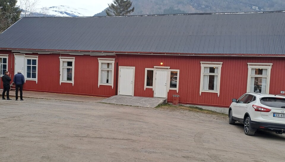 Et rødt hus med hvite detaljer. Foran huset står en hvit bil parkert og to mennesker