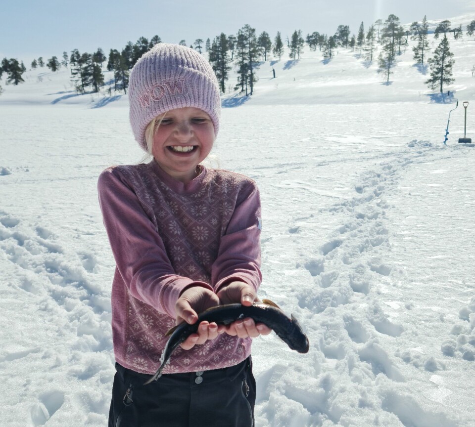 Ei jente som står i snøen og holder en liten fisk. I bakgrunnen et fiskehull i isen.