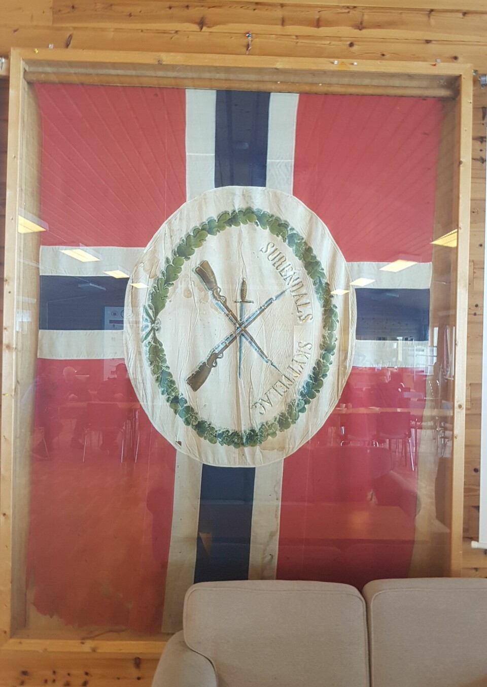 En fane ser ut som det norske flagget med en logo hvor en krans omringer to gevær i krys over hverandre