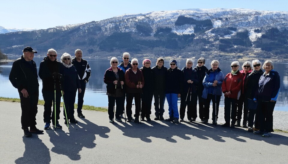 Ca 20 fritidskledde personer som står oppstilt ved sjøen. Det er sol, og snøkledde fjell i bakgrunnen.