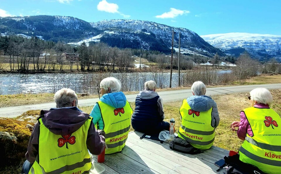 Fem damer med grått hår, fire av dem med refleksvester merka med sanitesforeninga sin logo og 'Kløvertur', sitter med ryggen til og ser mot elva og snøkledde fjell.