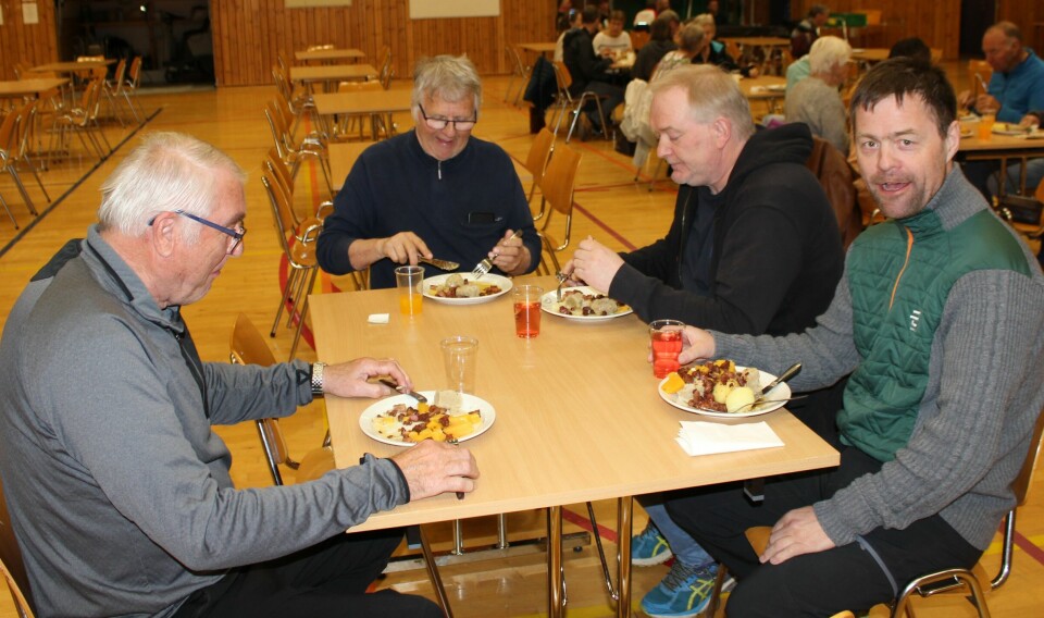 Fire menn sitter rundt et bord med hver sin middagstallerken og spiser.