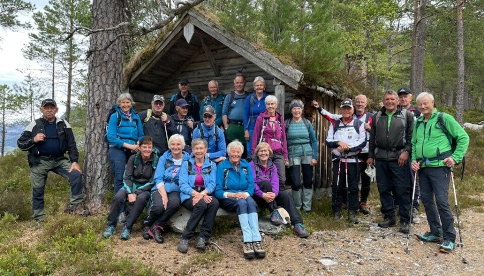 Mange mennesker i turklær står og sitter foran et trehus i skogen