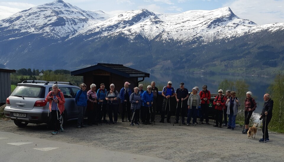 Ei gruppe pensjonister som står oppstilt på en parkeringsplass i forbindelse med tur. Snøkledde fjell i bakgrunnen.