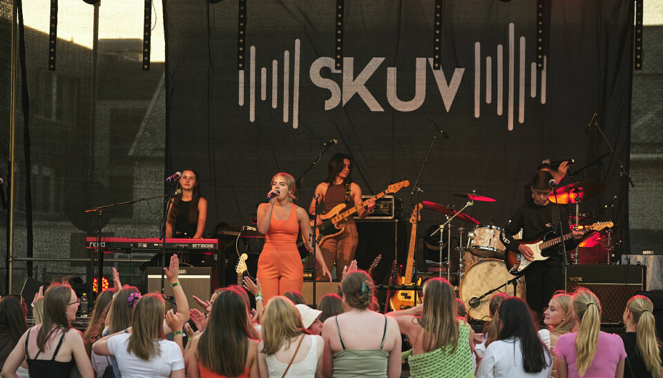 En scene med et band og en kvinnelig vokalist. Foran scenen står mange unge tilskuere