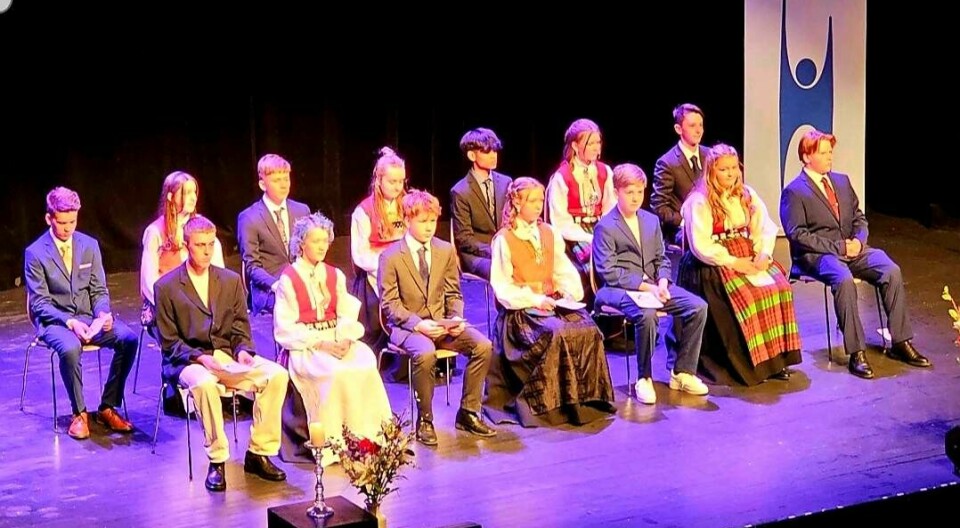14 konfirmanter i dress og bunader sitter på hver sin stol på en scene. Bak står en plakat med human-etisk forbund sin logo.