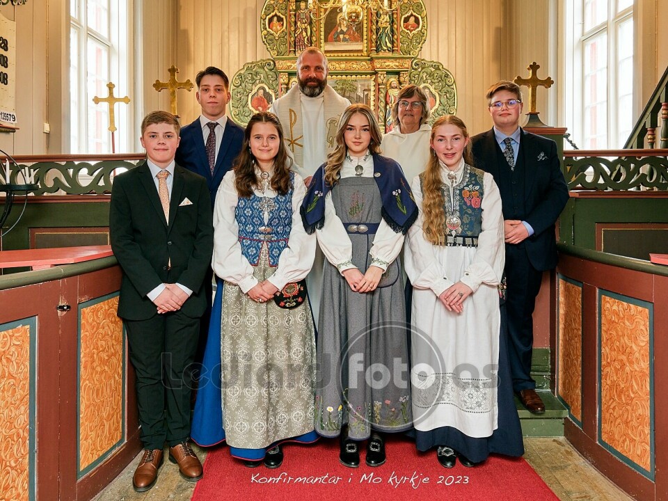 Seks konfirmanter i dress og bunader oppstilt foran alteret i kirka. Bak dem står en prest.