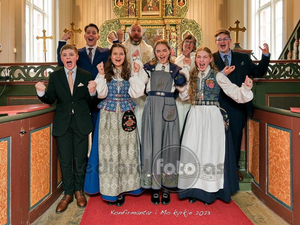 Seks konfirmanter i dress og bunader oppstilt foran alteret i kirka. De jubler og løfter hendene i været. Bak dem står en prest og ei dame i kirkekappe.