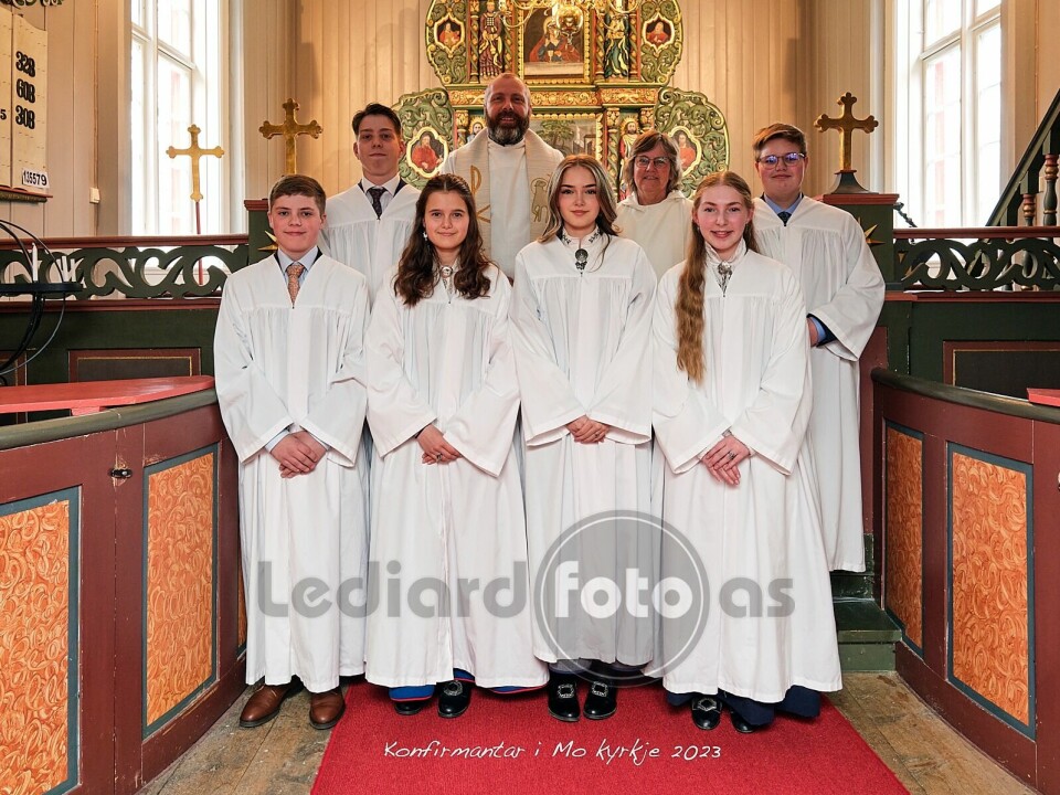 Seks konfirmanter i hvite konfirmantkapper oppstilt foran alteret i kirka. Bak dem står en prest og ei dame i kirkekappe.