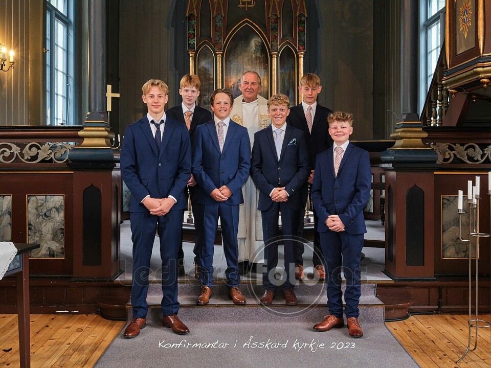 6 konfirmanter, alle gutter kledd i dress, oppstilt foran alteret i kirka. Bak dem står en prest.